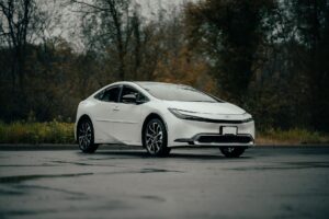 Modern white car parked on a wet road with autumn trees in the background.