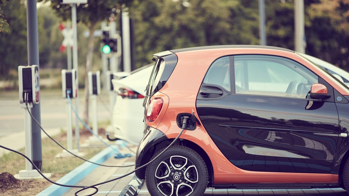 Petite voiture électrique orange et noire branchée à une borne de recharge sur une place de stationnement, avec d'autres bornes et une voiture blanche en arrière-plan.