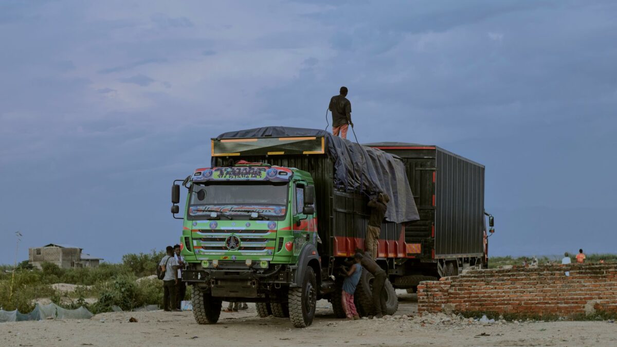 Camion coloré décoré de motifs traditionnels, stationné sur terrain sablonneux, personnes chargeant/déchargeant remorque, ciel couvert, constructions rudimentaires en arrière-plan.