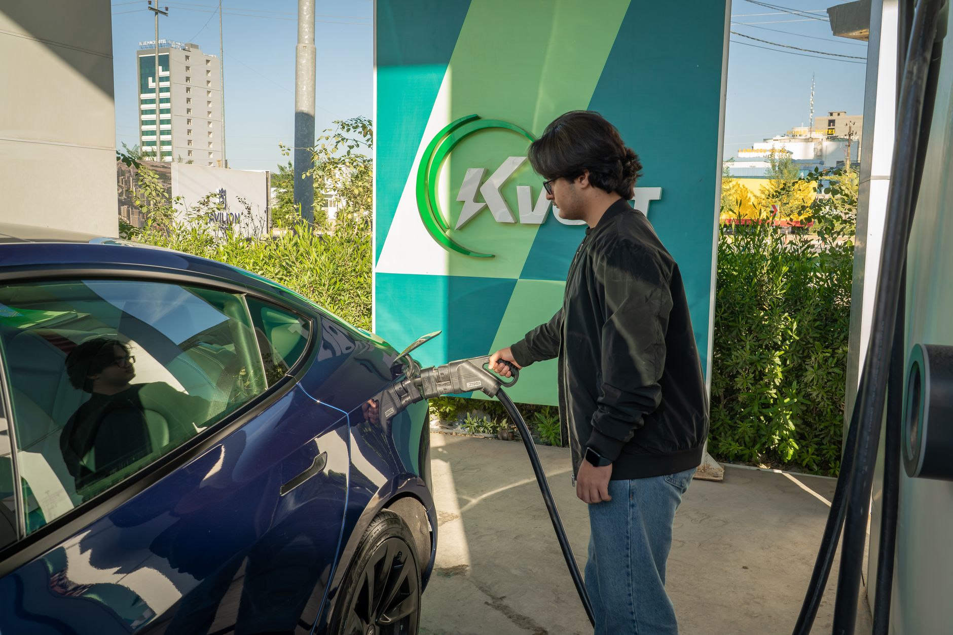 A person plugging a charging cable into the side port of a blue electric vehicle at a charging station.