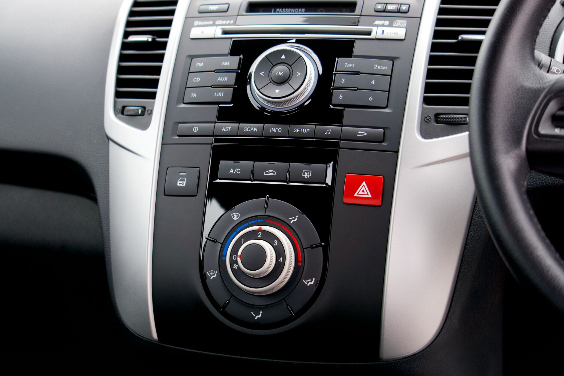 Car center console with radio buttons, a rotary controller, a climate dial, a red hazard button and air vents, with part of the steering wheel at right.