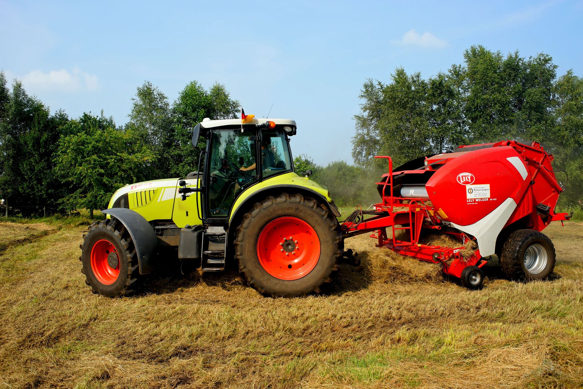 A green tractor pulling a red round baler across a mown hay field, with trees and a blue sky in the background.
