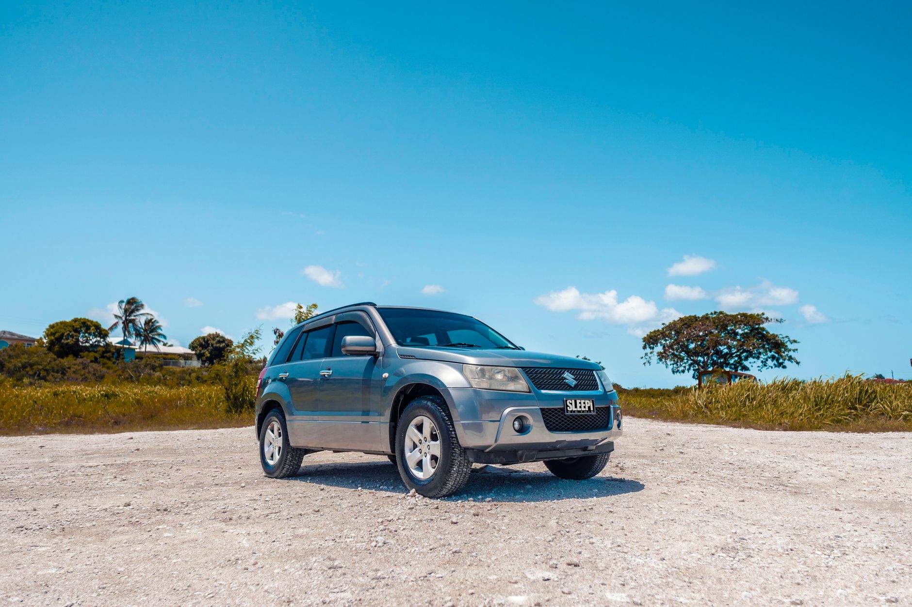 A grey compact SUV parked on a light-colored gravel surface under a bright blue sky.