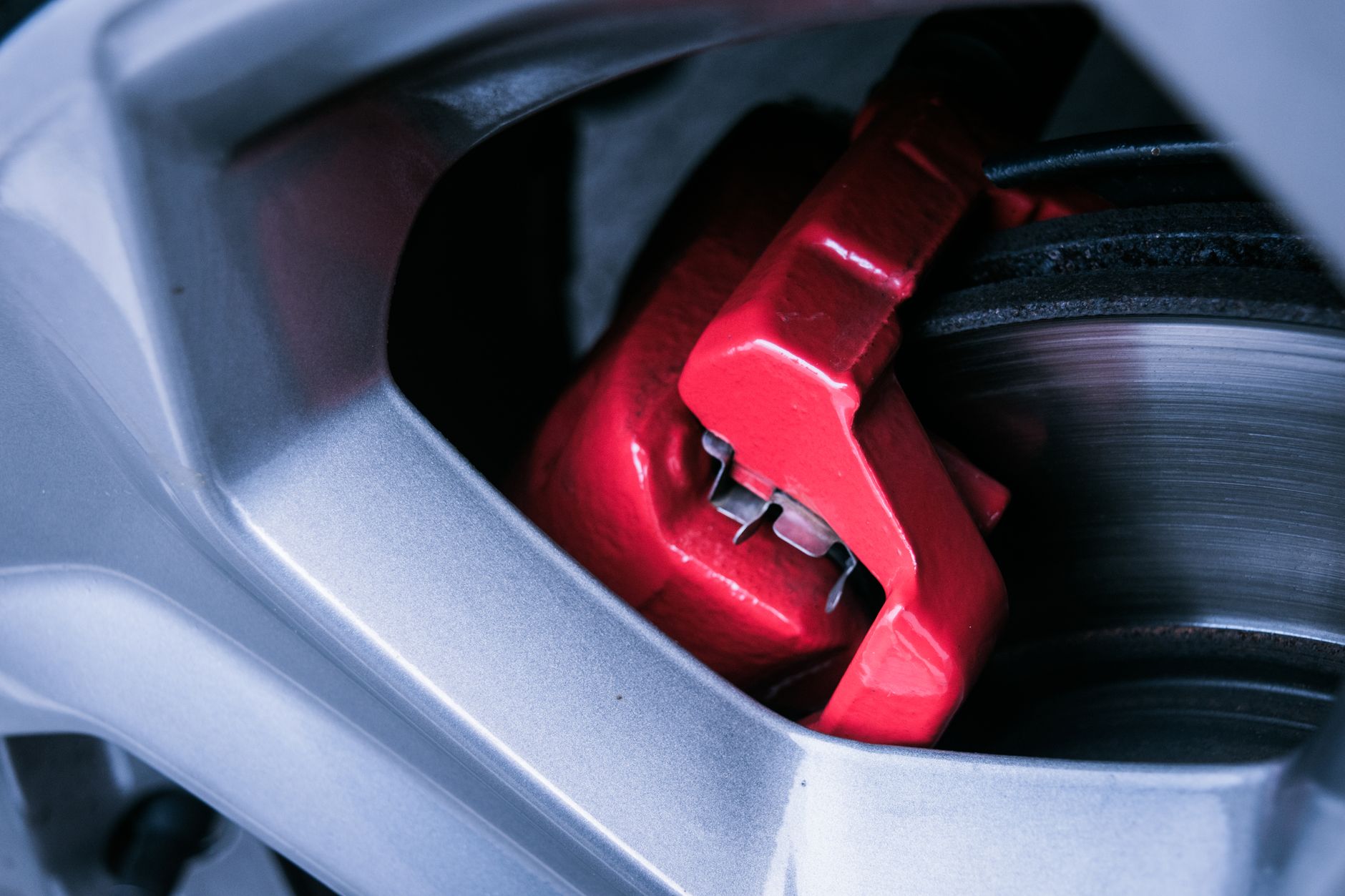 Red brake caliper behind a silver alloy wheel spoke, next to a brake disc, in close-up.