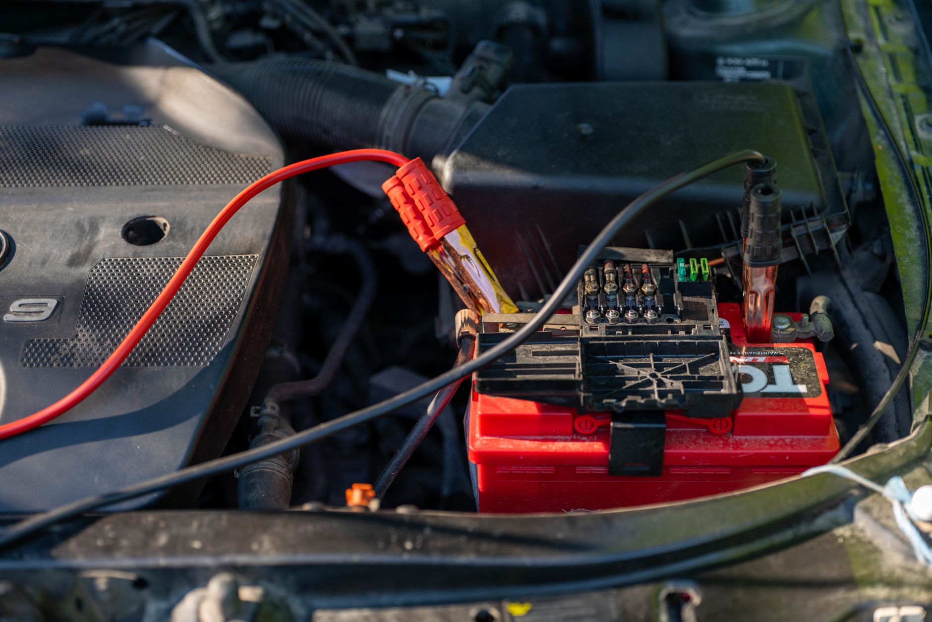 Red car battery in an engine bay with jumper cables attached to its terminals, beside a black engine cover.